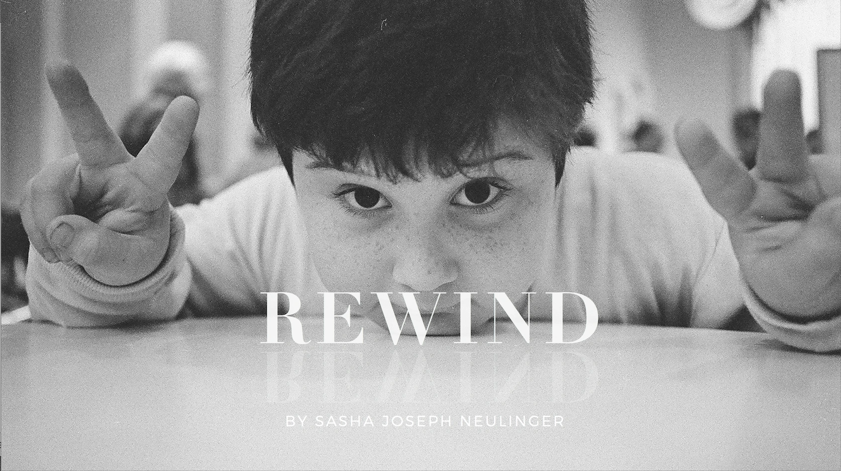 A black-and-white photo of a young boy looking into the camera, holding up two peace signs. The word REWIND and By Sasha Joseph Neulinger are overlaid on the image.