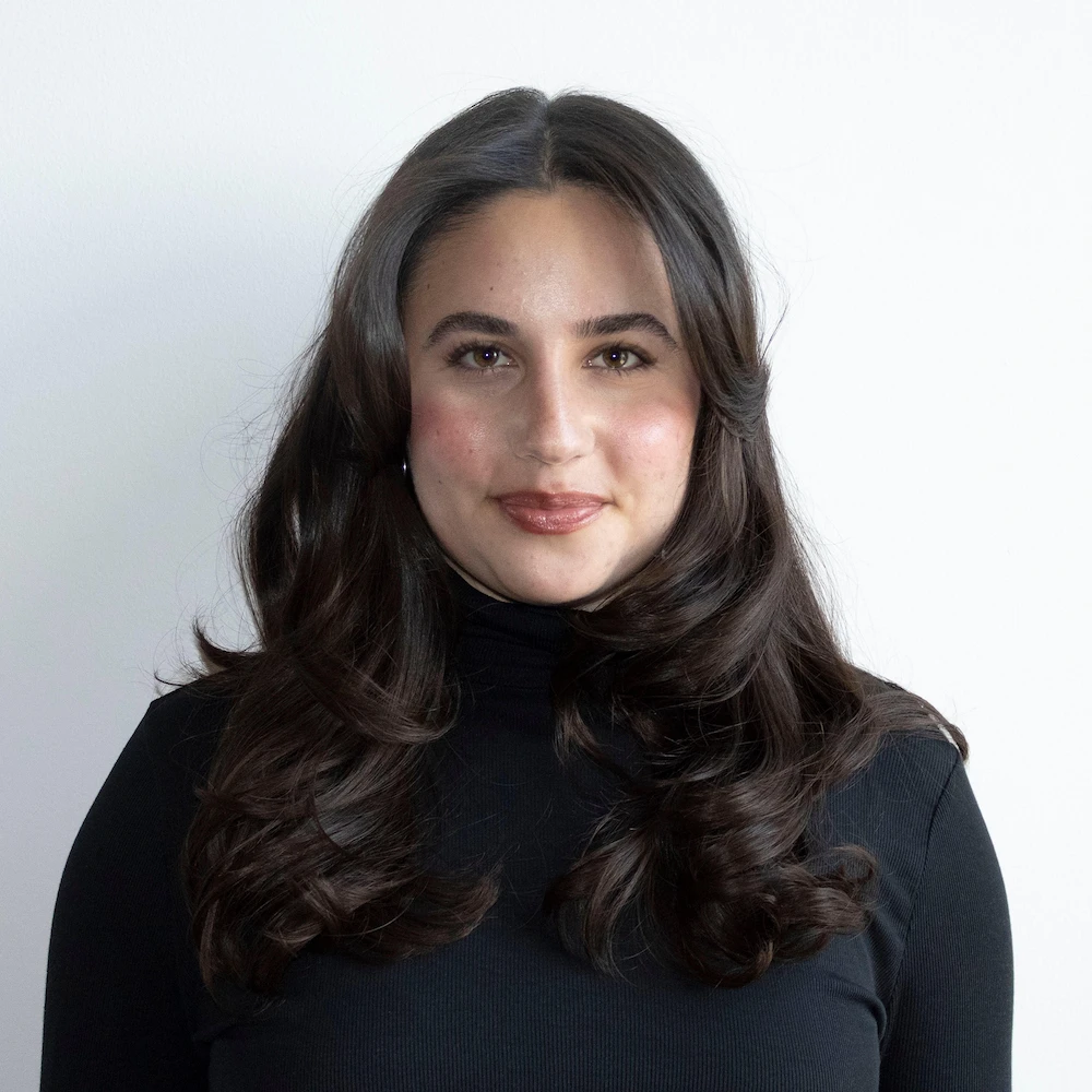 Jenna Lotterman Headshot-1Kx1K A person with long, wavy dark hair wearing a black turtleneck top stands in front of a plain white background, smiling softly at the camera.
